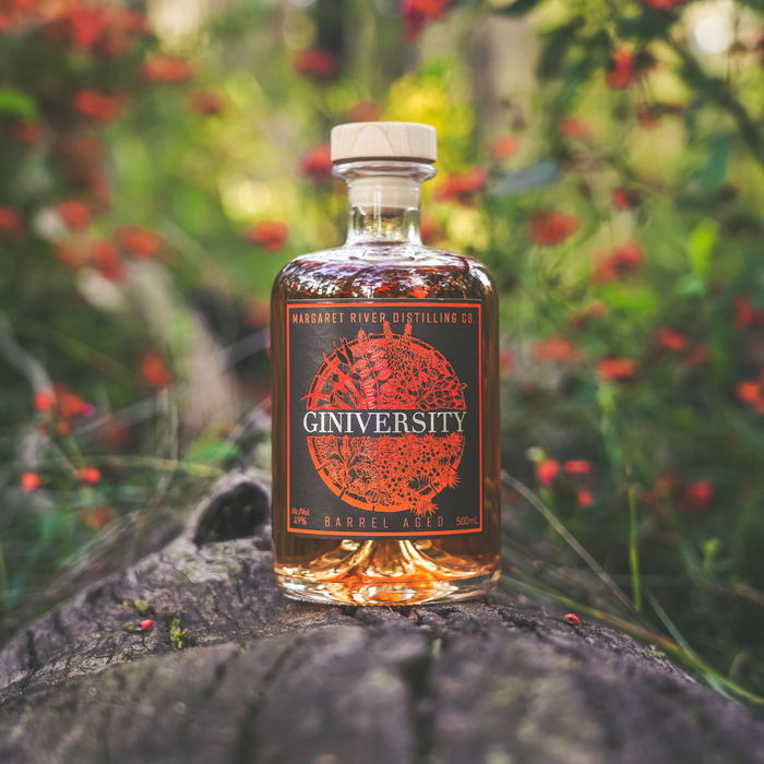 Giniversity Barrel Aged 500ml bottel on a log with red flowers on the background